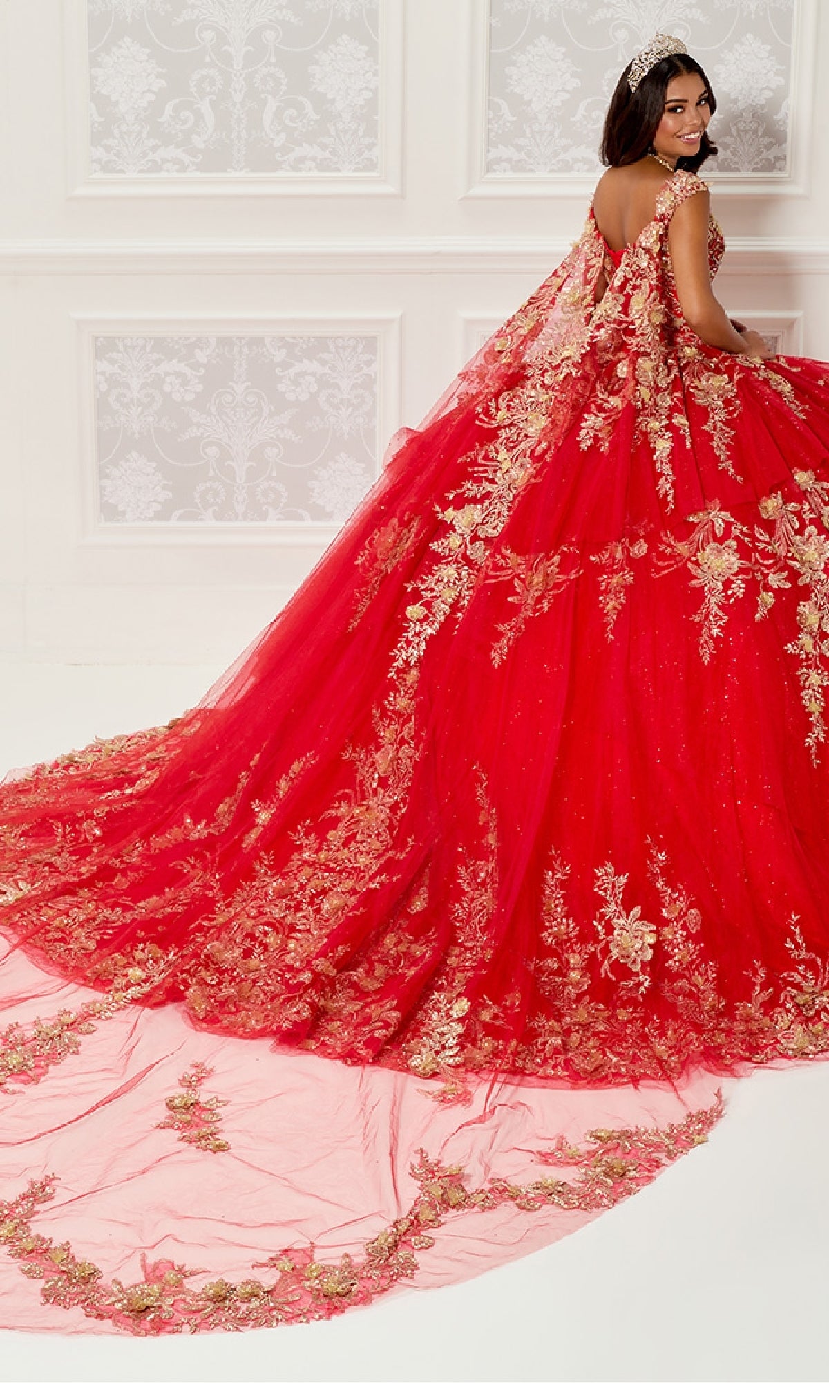 A woman wears the Quinceanera Dress PR30119 By Princesa, featuring gold embroidery and a glitter tulle gown with a long train, standing in an ornate room as her detachable cathedral cape flows elegantly behind her.